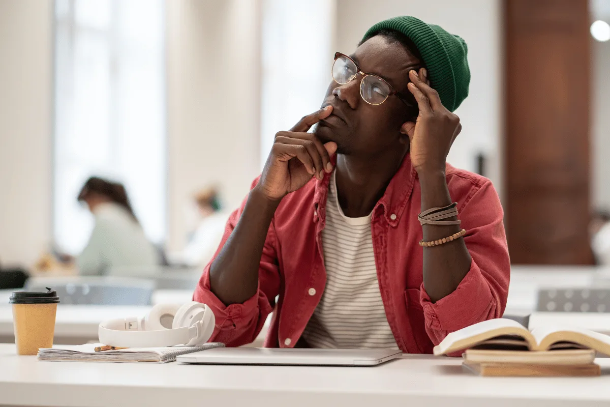 Un étudiant s'interrompt, la main sur la tempe, devant son ordinateur fermé et un livre ouvert.